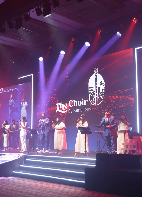 A wide shot of a brightly lit stage at a Motilal Oswal event featuring "The Choir by Sampoorna." A group of seven vocalists in formal blue suits and elegant white dresses perform under dramatic purple and red spotlights. A large digital screen in the background displays the event logo and vibrant abstract graphics, while professional audio equipment and stage lighting surround the performers.